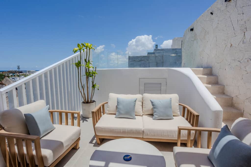 Outdoor balcony with white cushioned seating, potted plant, and views of blue sky, clouds, and surrounding rooftops at Luna in Playa del Carmen.