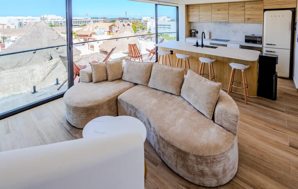 Living room with large glass windows overlooking rooftops and sky in Playa del Carmen, featuring a beige sectional sofa and modern kitchen at Luna.