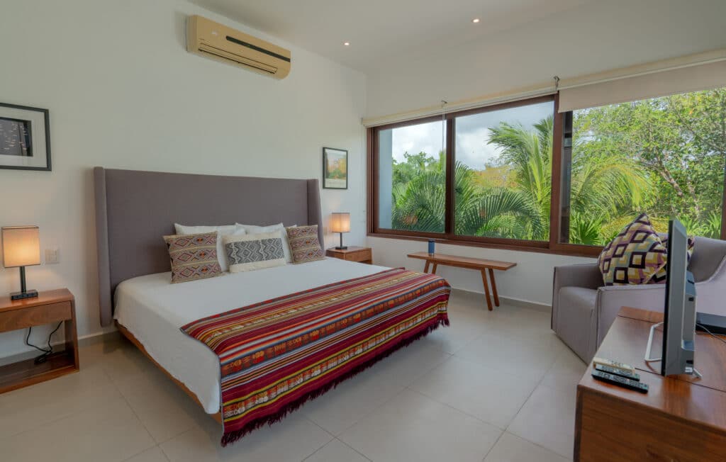 Interior of a bedroom with a large window showing tropical greenery, featuring a bed with colorful striped bedding and wooden furniture in Akumal.