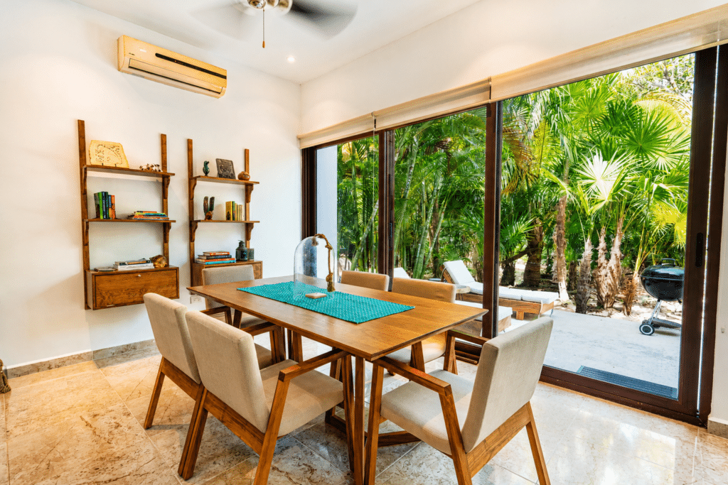 Dining room with six beige chairs around a wooden table, large sliding glass door leads to a lush tropical outdoor area in Akumal.