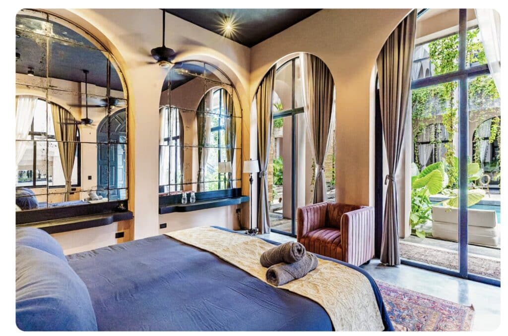 Interior of a bedroom with large arched windows, beige curtains, a bed with rolled towels, and a view of a lush outdoor courtyard in Tulum.