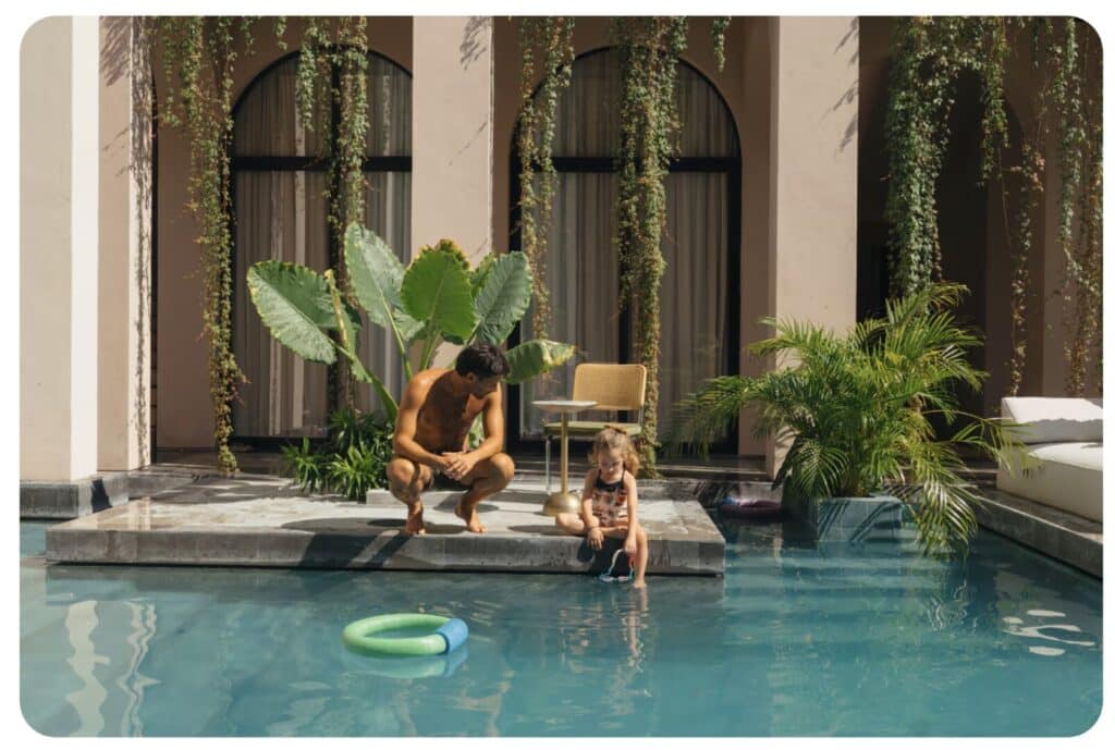 A man and a young girl sitting by a swimming pool with lush green plants and large arched windows in the background in Tulum.