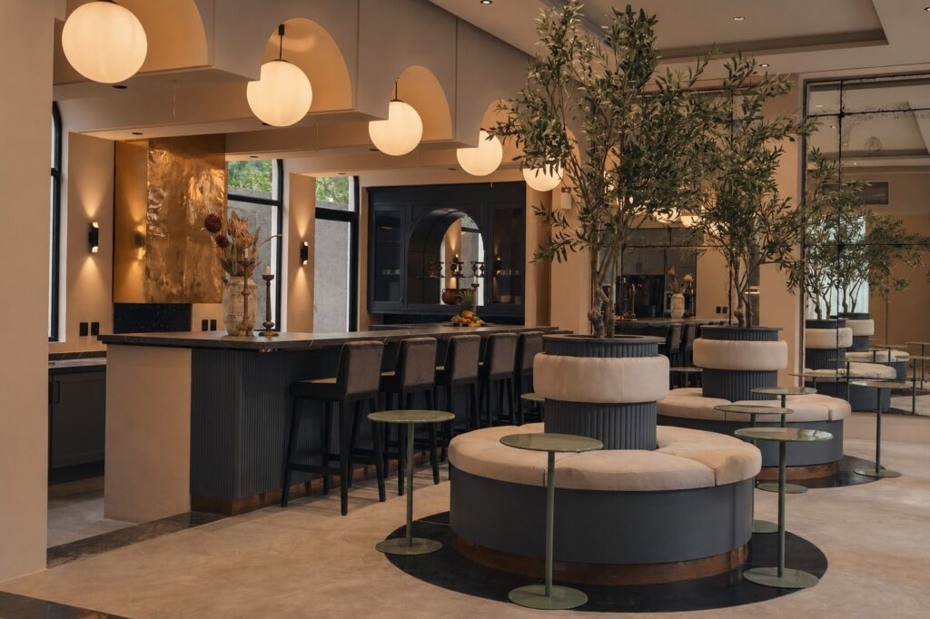 Interior of a modern lounge with large potted trees, curved seating, bar area, and warm lighting in Tulum.