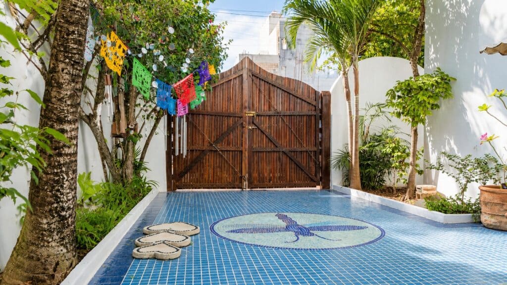 Private backyard with a blue tile mosaic floor, lush tropical plants, colorful papel picado banners, and a wooden gate in Playa del Carmen.