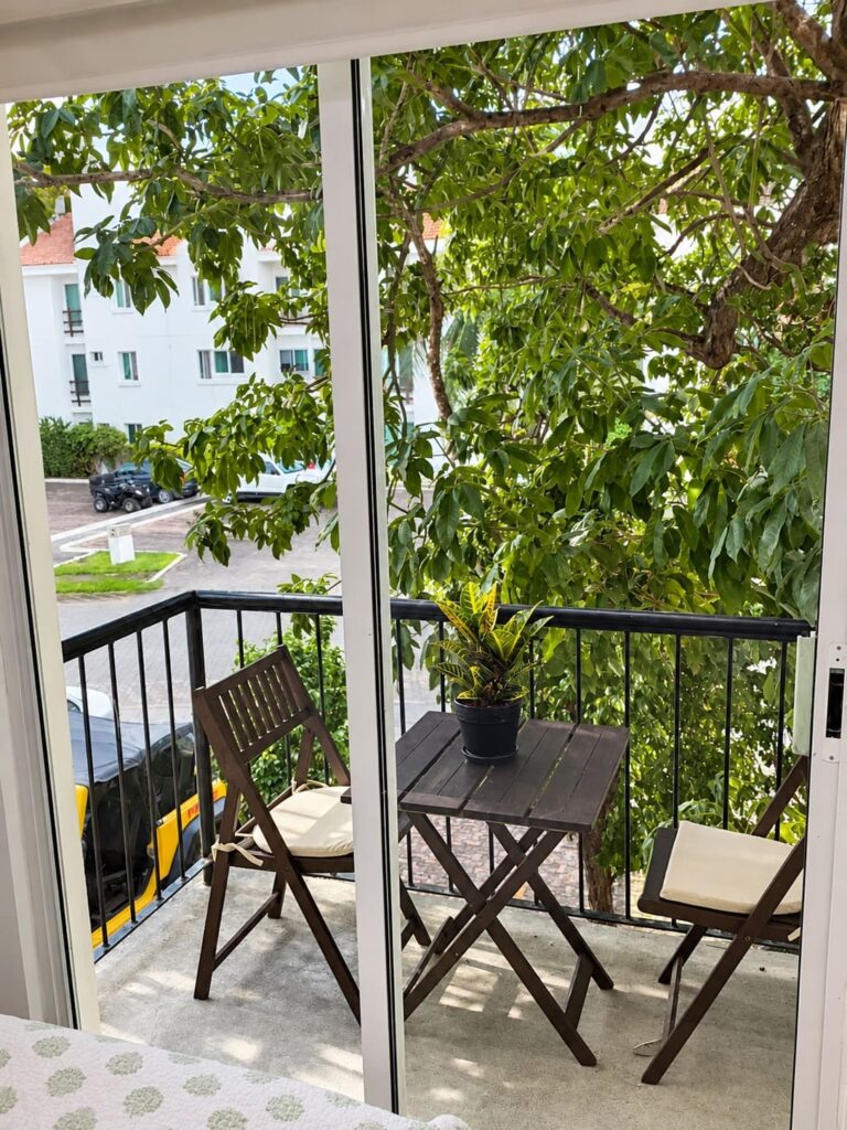 View of a small balcony with wooden table and chairs, overlooking a tree and nearby residential buildings in Playa del Carmen.