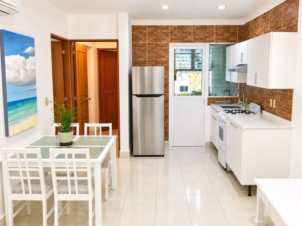 Kitchen with white cabinets, a stainless steel refrigerator, brick backsplash, and a window overlooking a garden in Playa del Carmen.