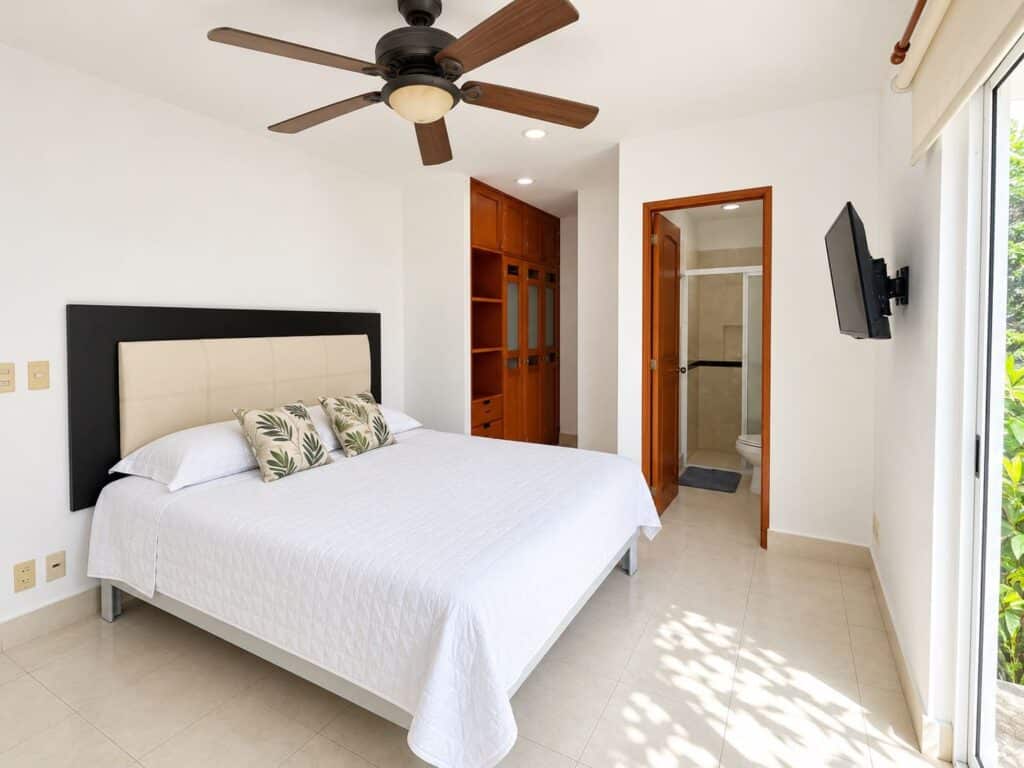 Bedroom with white bedding, wooden ceiling fan, sliding glass door, and view of lush greenery in Playa del Carmen.