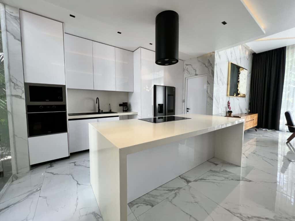 Modern kitchen with white cabinetry, black appliances, and a marble floor, adjacent to a living area with a wooden console and dark curtains at Club Real in Playa del Carmen.