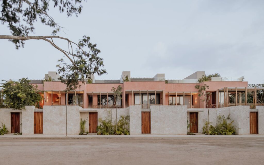 Modern pink and beige multi-unit residential building with trees in front, set against a cloudy sky at Entorno Tulum Villas.